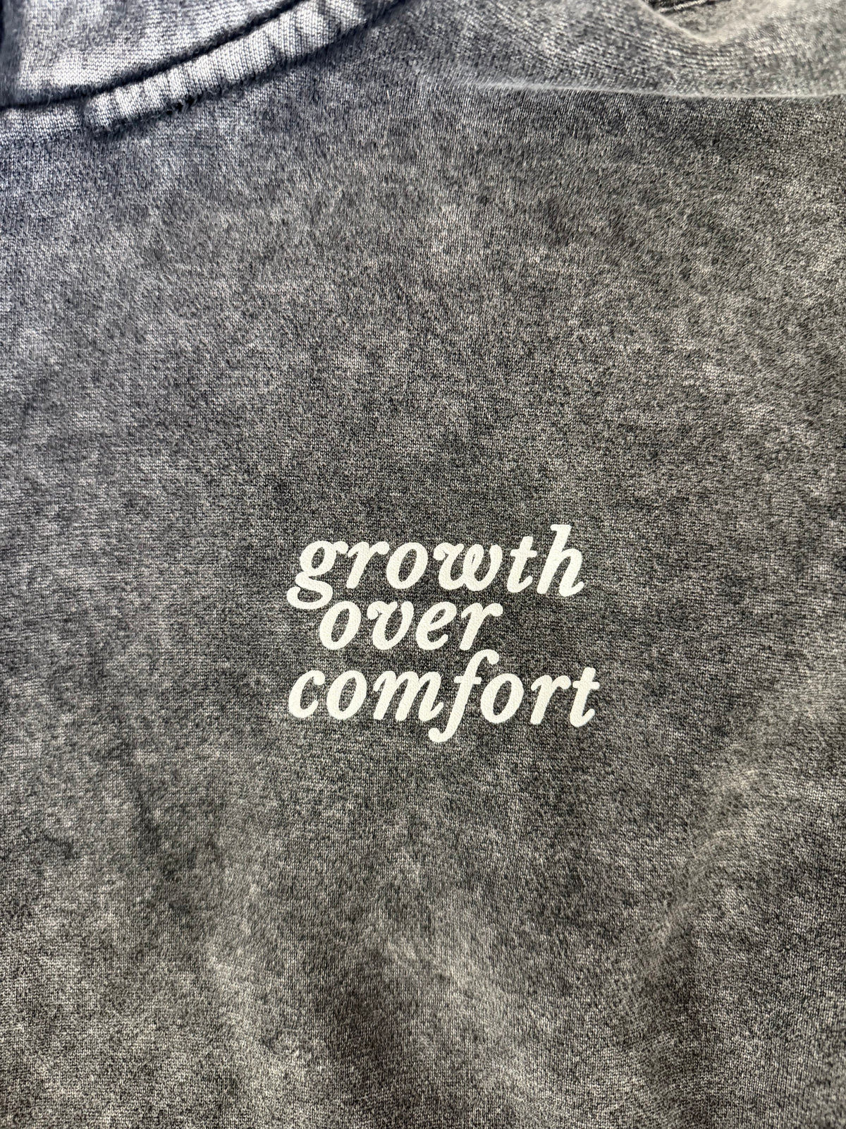 Close-up of a dark gray mineral-wash crewneck sweatshirt featuring the phrase 'growth over comfort' printed in white script across the chest.